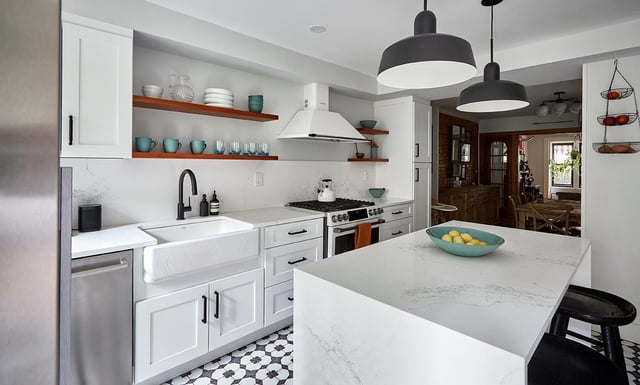 A modern kitchen features white cabinets with black hardware, an undermount white farmhouse sink with a black faucet, white marble-look countertops, open wooden shelving above the counters, and a white kitchen island with a bowl of lemons, all set atop a black and white patterned tile floor, with a dining area visible through a doorway in the background.