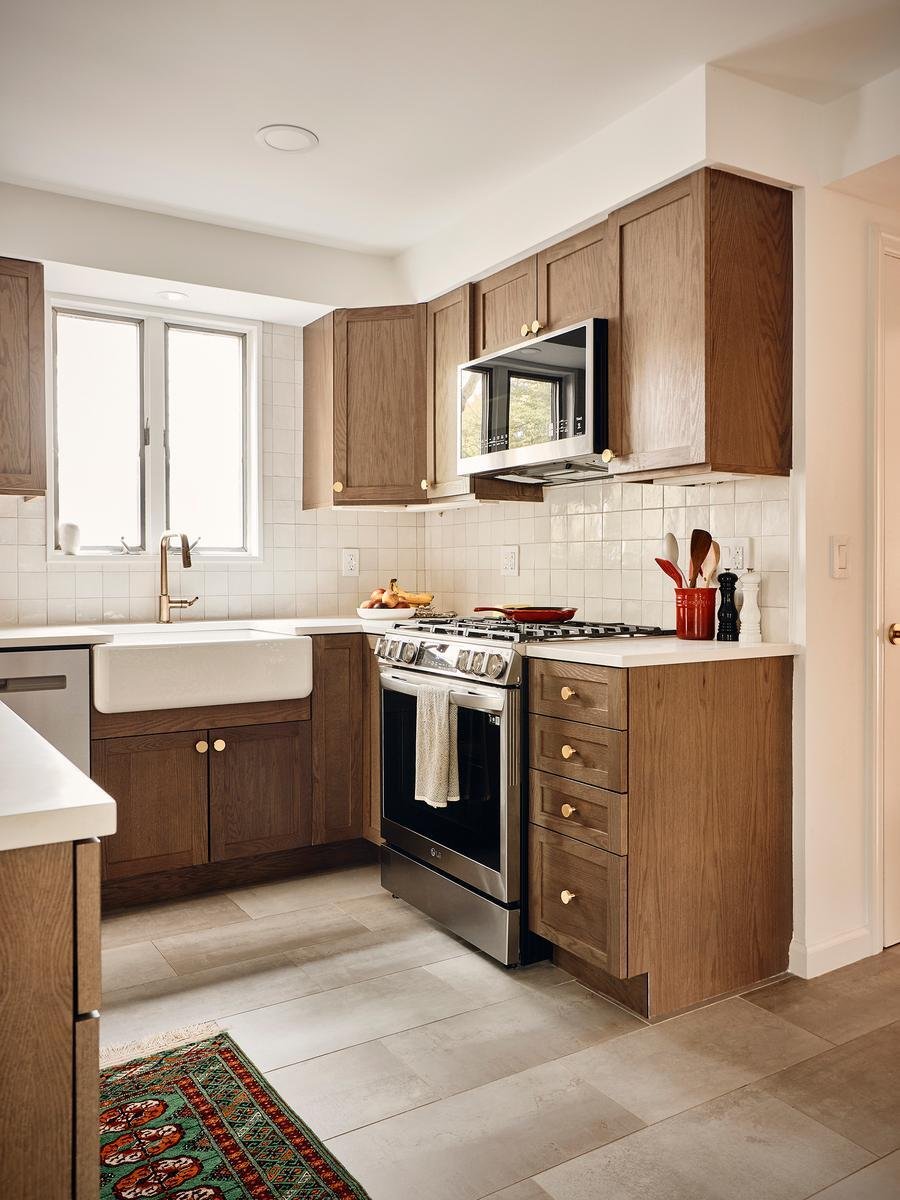 The image shows a modern kitchen with natural wood cabinetry, white square tile backsplash, white countertops, and stainless steel appliances, including a range and over-the-range microwave.