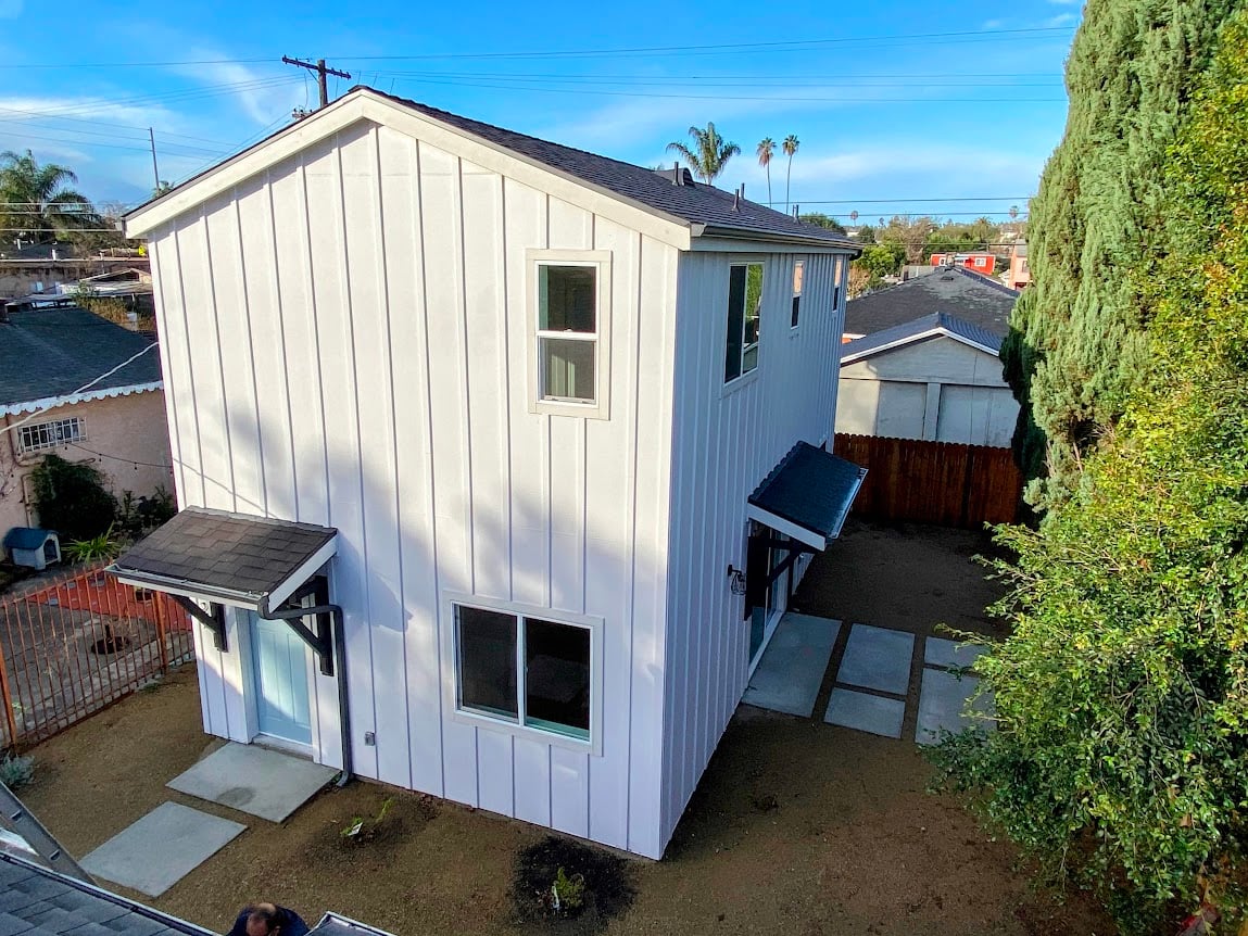  A two-story, modern farmhouse-style ADU featuring crisp white board-and-batten siding, a light blue front door, and a minimalist gravel yard with stone pavers.