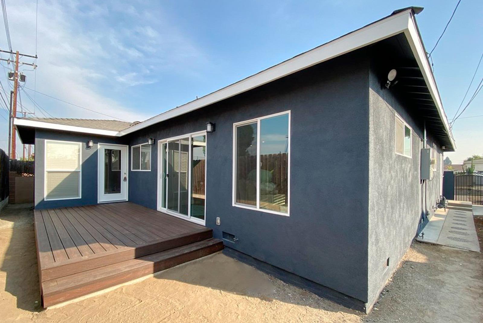  A contemporary single-story home featuring dark charcoal grey stucco walls, crisp white window trim, and a spacious raised wooden deck leading to sliding glass doors.