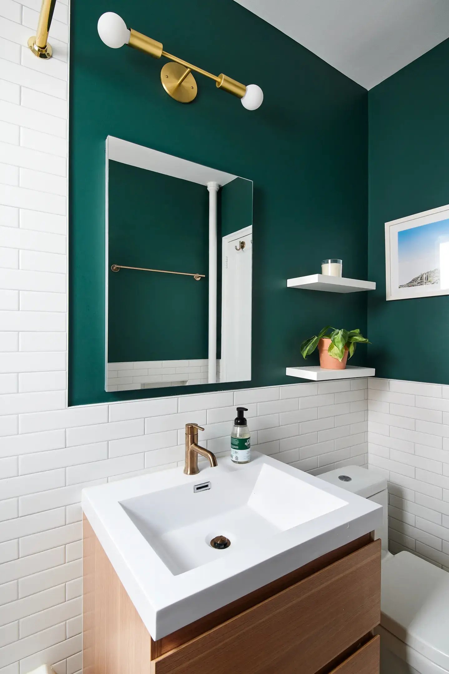 A modern bathroom features a light wood vanity with a white integrated sink and gold faucet, set against a wall with white subway tile wainscoting and a dark green upper portion, which holds a rectangular mirror with a thin white frame, a gold two-bulb vanity light, a gold towel bar, and two white floating shelves.