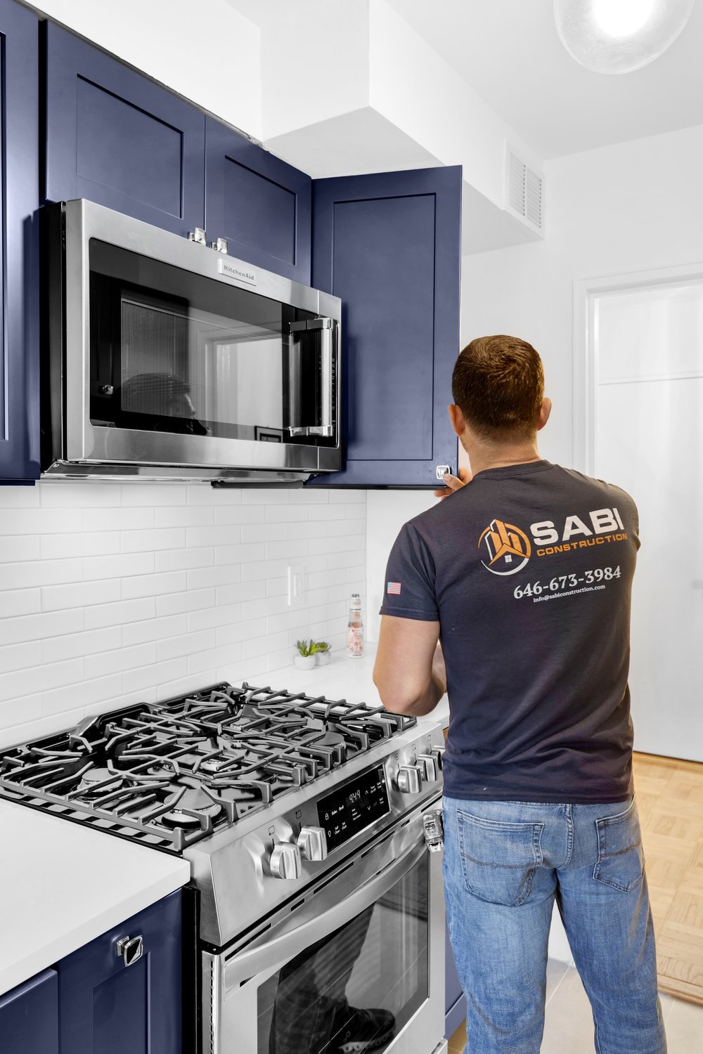 A Sabi Construction worker in a navy blue shirt and jeans is installing hardware on a dark blue kitchen cabinet above a stainless steel range and microwave.