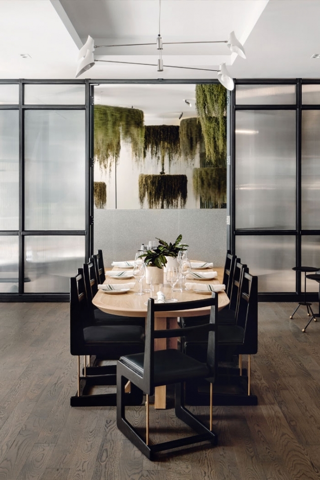 A private dining area features a light wood oval table set with tableware and a plant centerpiece, surrounded by black, angular dining chairs with brass accents, all situated on a dark wood floor and framed by black-grid glass and translucent panels that open to reveal hanging moss installations in the background.