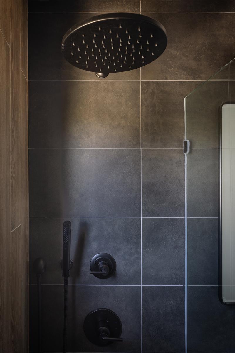Moody modern shower with a matte black rainfall showerhead and fixtures, dark charcoal stone-look tile walls with clean grout lines, frameless glass panel, and a minimalist spa-inspired design.
