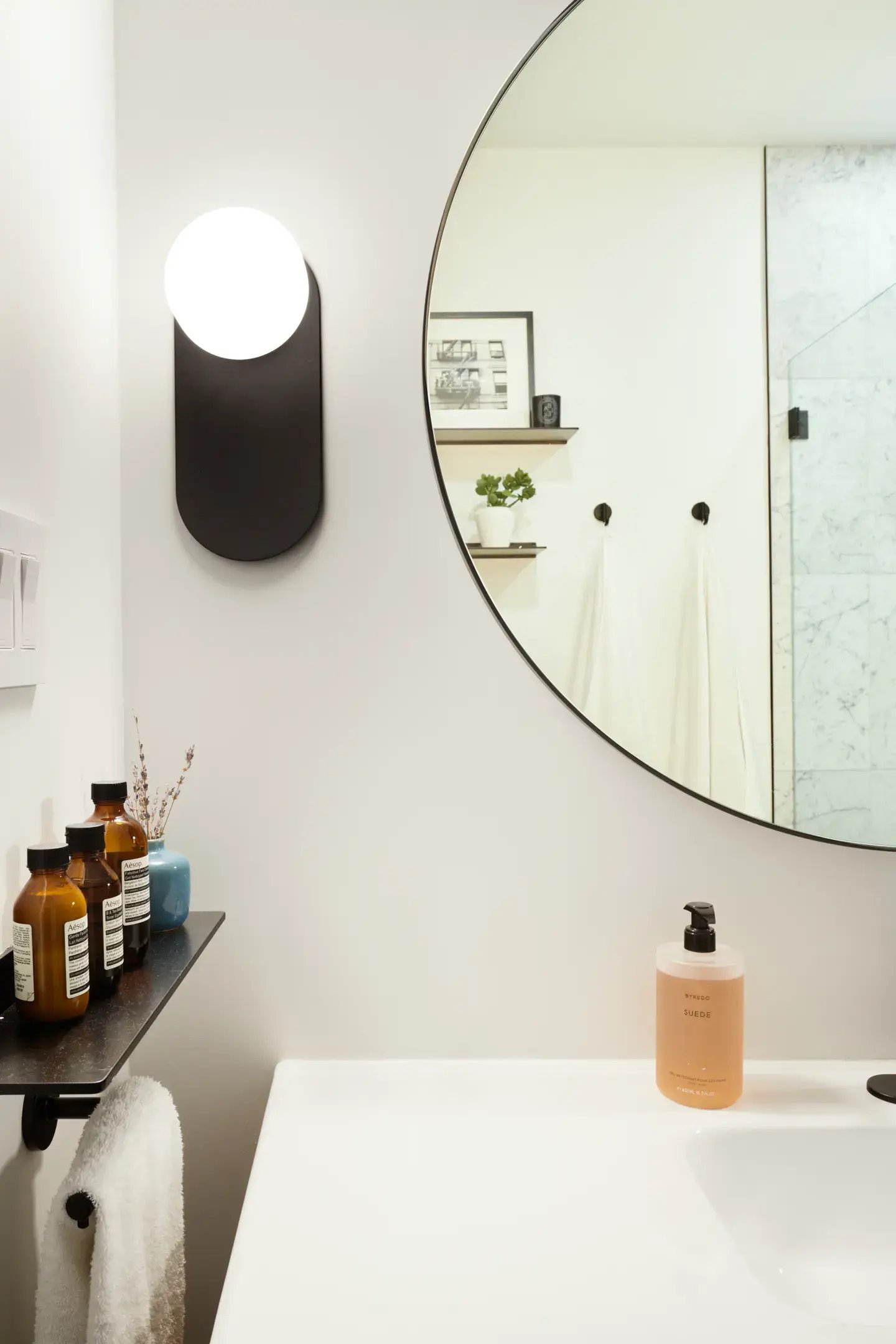 A modern white bathroom features a large circular mirror reflecting white walls, a black-and-white picture frame, a small shelf with a potted plant, and two black hooks with white towels, while the foreground shows a white sink with a pump bottle of liquid soap, a black sconce with a round white light, and a small black shelf holding several amber bottles.