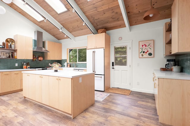 A bright kitchen featuring light wood cabinets, an island with a white countertop, stainless steel appliances, and a vaulted wood-plank ceiling with skylights.