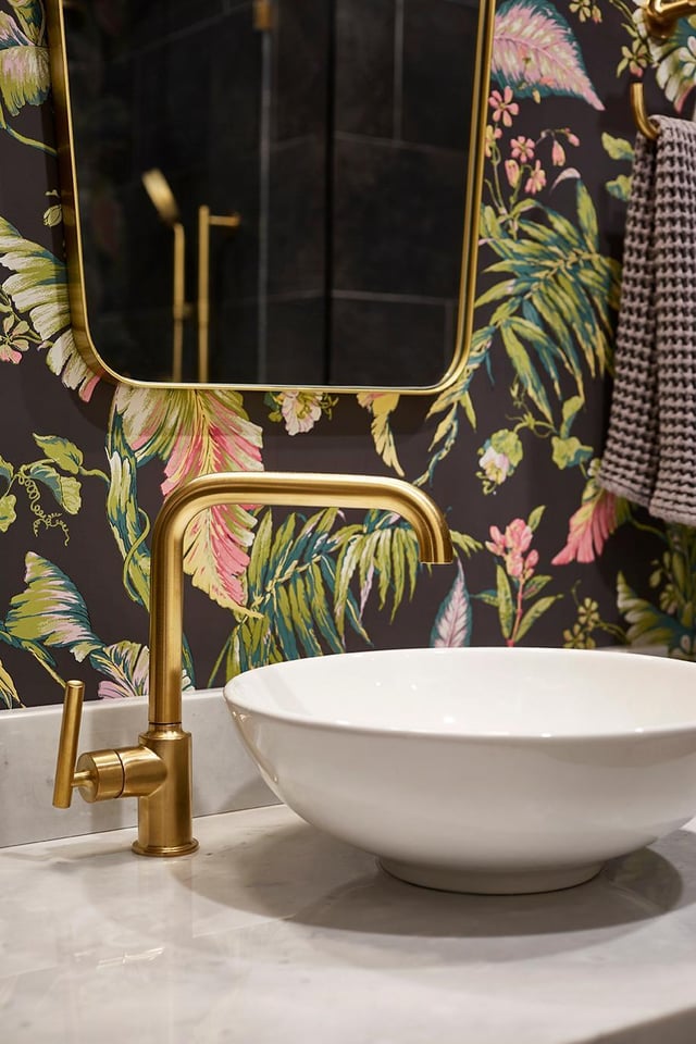 A white vessel sink and a tall gold-toned faucet sit on a white marble vanity counter against a dark wallpaper patterned with large pink and green tropical foliage, with a curved brass-framed mirror visible above.