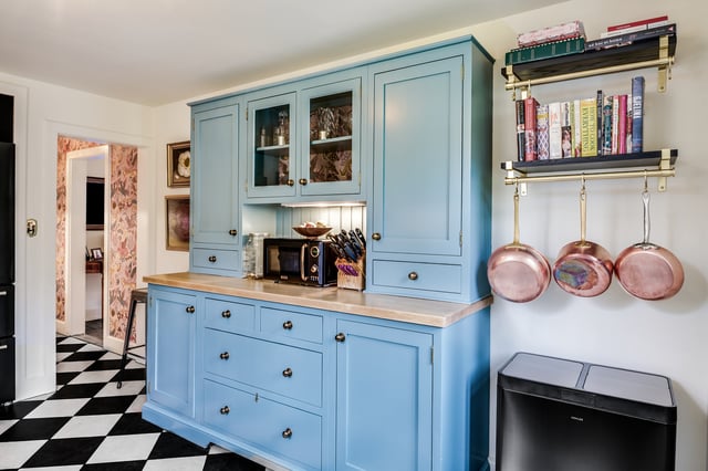 A kitchen features a large, light blue built-in hutch with glass-fronted upper cabinets, brass knobs, and a butcher block countertop, centered on a black and white checkered floor, with a black microwave and a knife block sitting on the counter, and two shelves to the right holding cookbooks and three copper pots hanging below them.