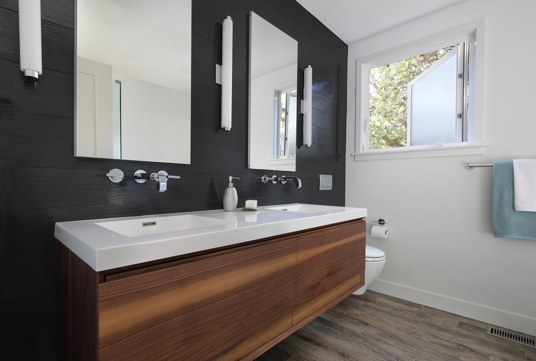 A sleek, high-contrast modern bathroom featuring a floating walnut double vanity, a textured black accent wall, and minimalist chrome fixtures under a bright window.