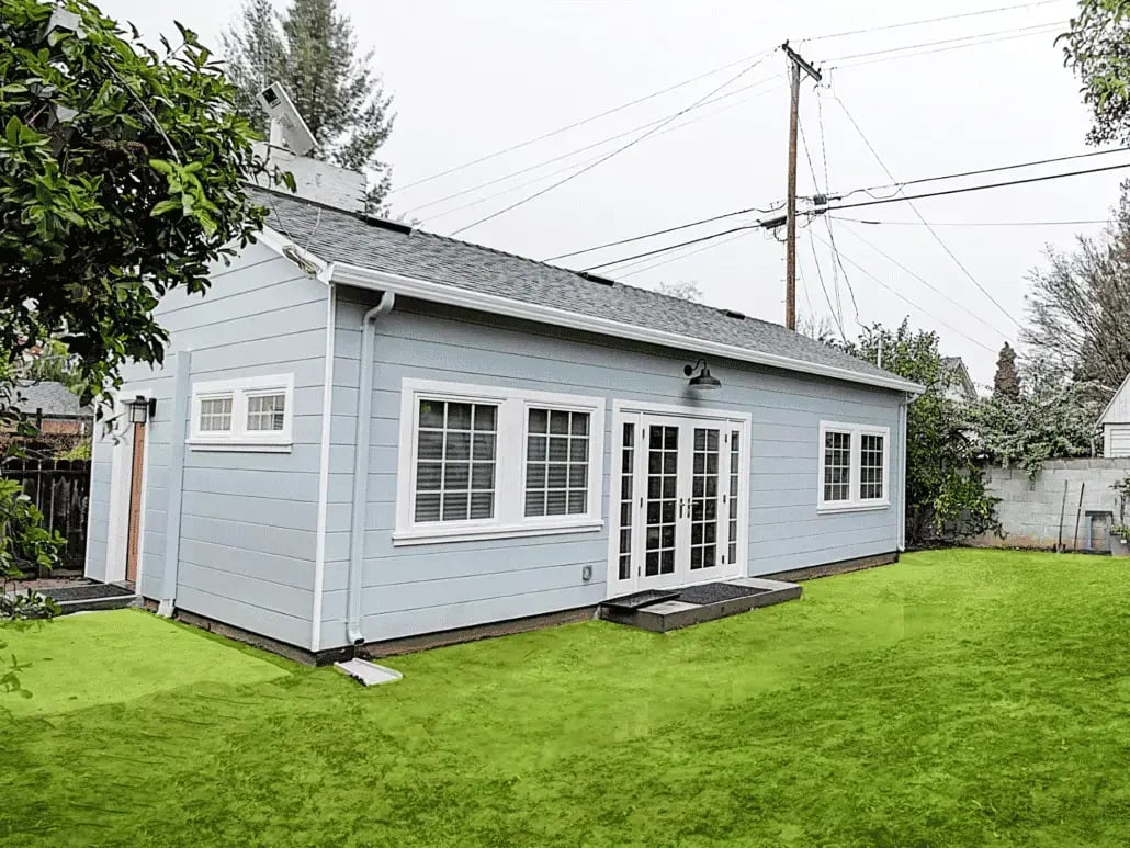  A charming, light blue cottage-style ADU featuring horizontal lap siding, crisp white trim, and elegant French doors that open onto a bright, manicured green lawn.