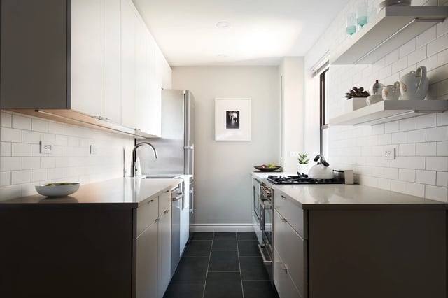 The image shows a bright, modern galley kitchen with white upper cabinets, gray lower cabinets, light gray countertops, white subway tile backsplash, a stainless steel refrigerator and range, and dark tiled flooring.