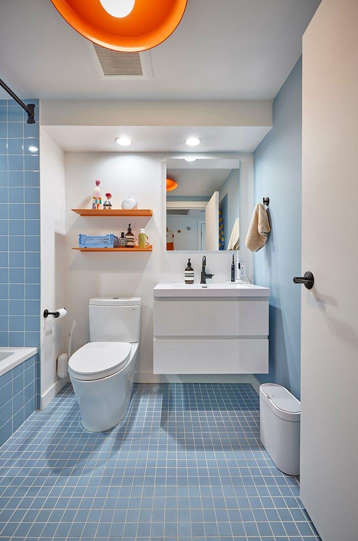 The image shows a contemporary bathroom with blue square-tiled walls and floors, a white floating vanity, a white toilet, and orange accent shelves, all illuminated by a unique orange light fixture above.