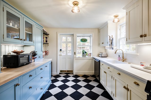 A kitchen features light blue lower cabinets with a wood countertop on the left and white upper cabinets, as well as white lower and upper cabinets on the right, all accented with gold hardware, a white farmhouse sink, and a black and white checkered floor, with a doorway and window at the back.