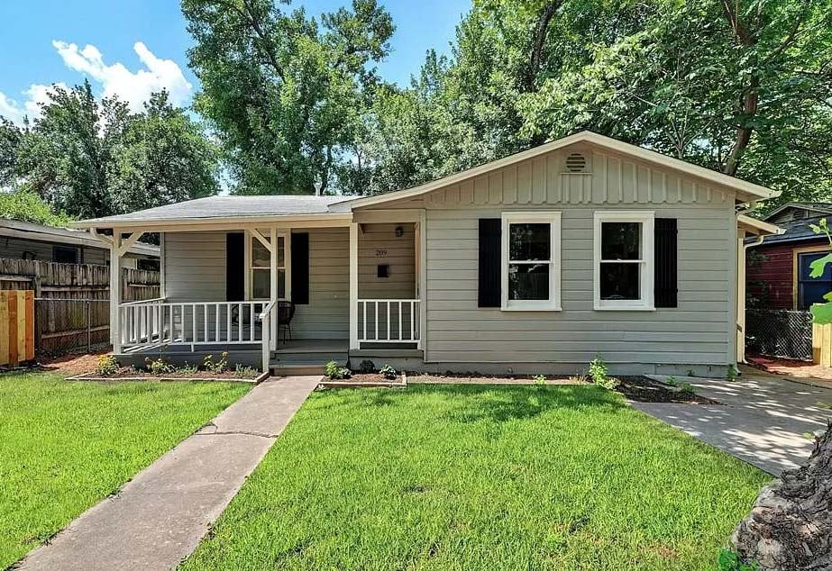 A cozy, traditional ranch-style home featuring light grey horizontal lap siding, dark black shutters, and a welcoming front porch with a white wooden railing overlooking a bright green lawn.