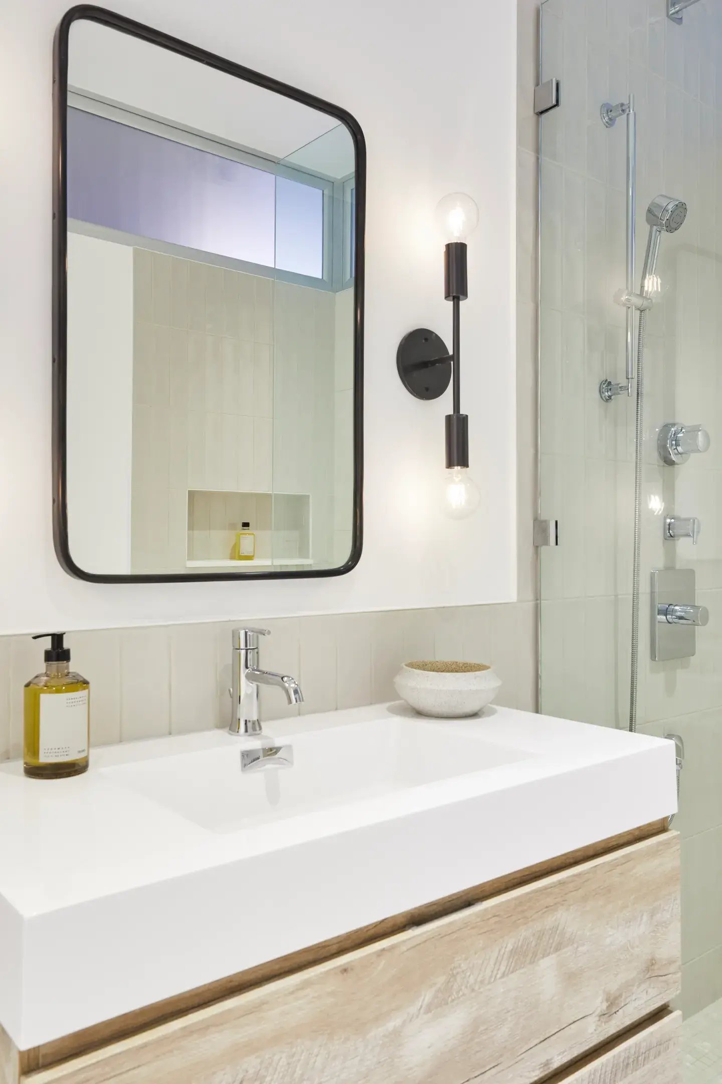 Bathroom vanity with a white rectangular sink, a wooden base, a black-framed mirror, and a black double-bulb light fixture on a wall with light tan subway tiles, next to a glass shower enclosure.