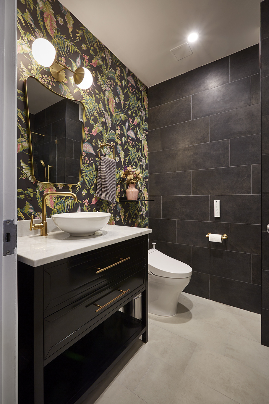 A remodeled bathroom in New York City with large format charcoal wall tile, large format taupe floor tiling, floral patterned wallpaper, brass fixtures, and a black vanity with marble countertop and vessel sink.