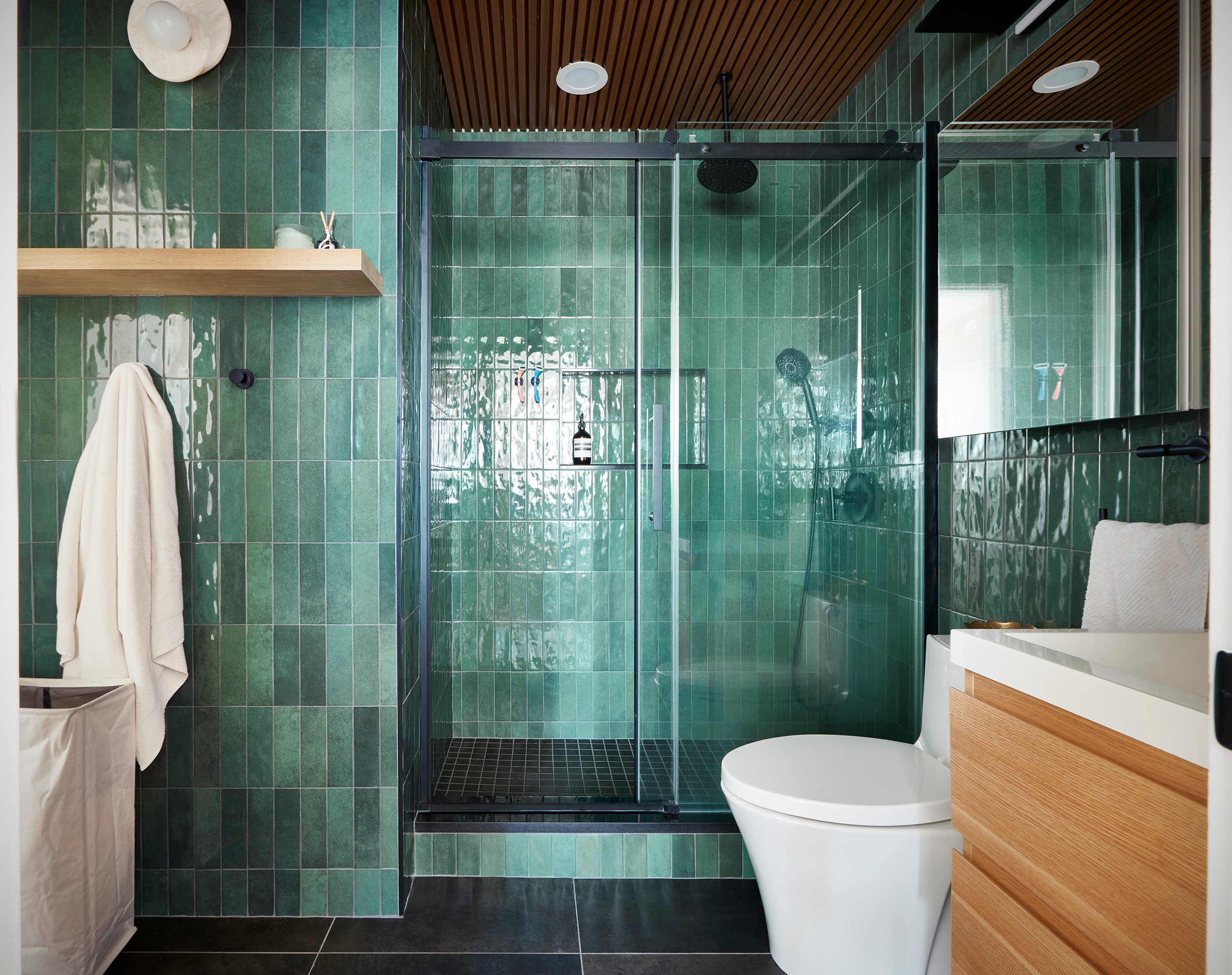Jersey City bathroom remodel with green wall tile, black large format floor tile, a glass sliding shower door, natural wood vanity, floating shelf, and matte black hardware.