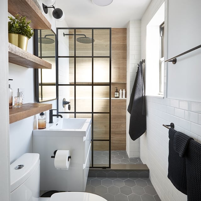 New York City bathroom with walk-in curbed shower with framed glass enclosure, wood grain wall tiling, shower niche, matte black fixtures, white floating vanity, gray hexagonal floor tile, and wood floating shelves.