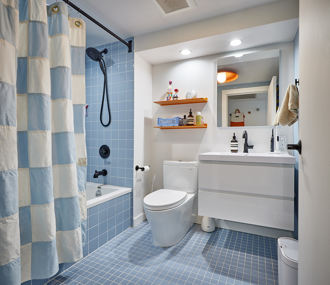 Brooklyn, New York City remodeled bathroom featuring blue wall and floor tiles, a tub-shower combo with matte black fixtures, checkered shower curtain, a floating white vanity, and orange floating shelves for storage.