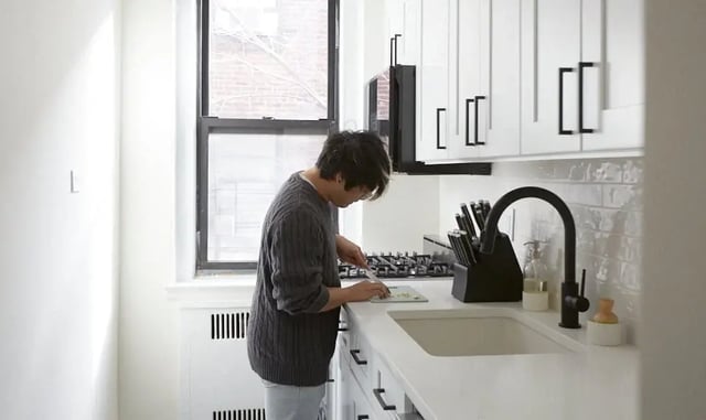 A modern, minimalist galley kitchen featuring white Shaker-style cabinetry with matte black hardware, a sleek white countertop with an integrated undermount sink, and a black gooseneck faucet.
