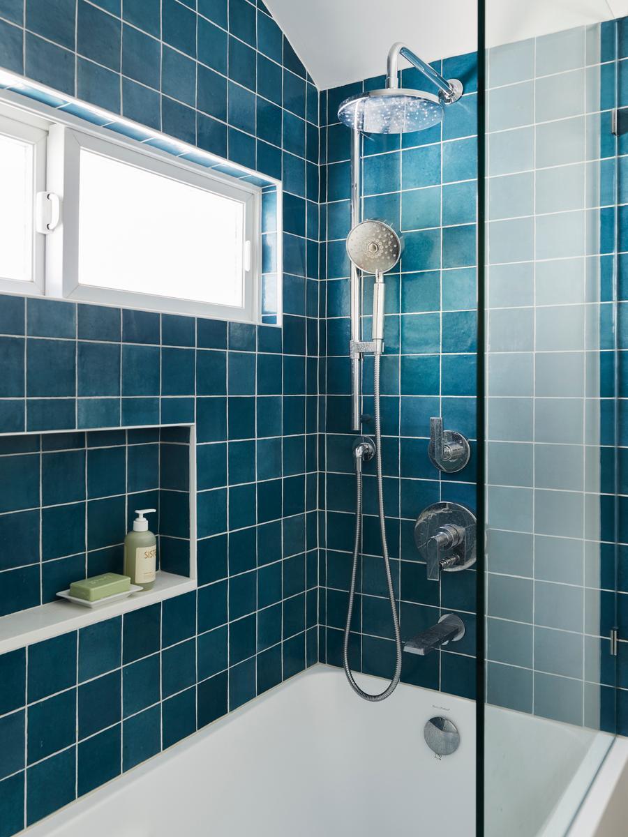 Blue-tiled shower with white grout lines, featuring a chrome ceiling-mounted rain showerhead, adjustable handheld sprayer, built-in wall niche, glass partition, and small frosted window for natural light.