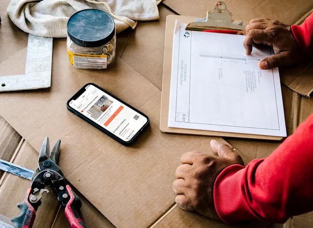 A worker reviews blueprints and a phone on a messy surface.