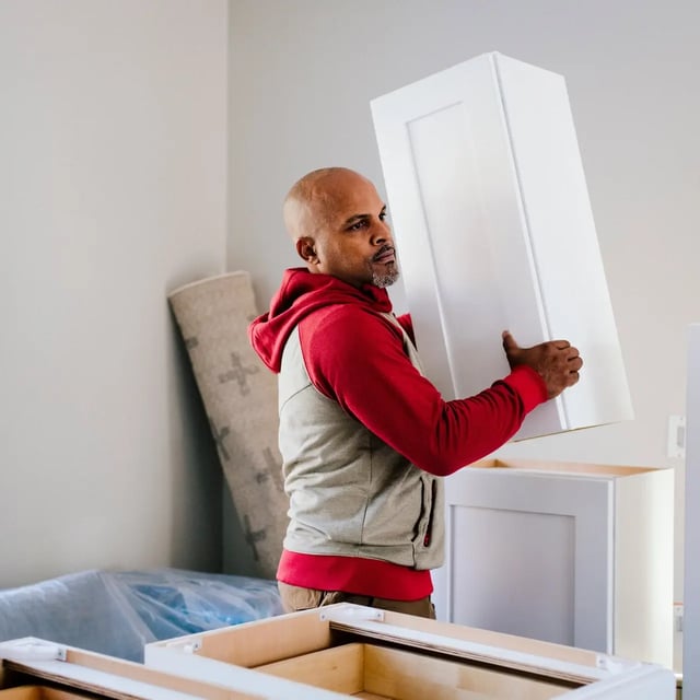 A man indoors lifting and positioning a white cabinet during a home remodeling project, with unfinished cabinetry and renovation materials visible around him.