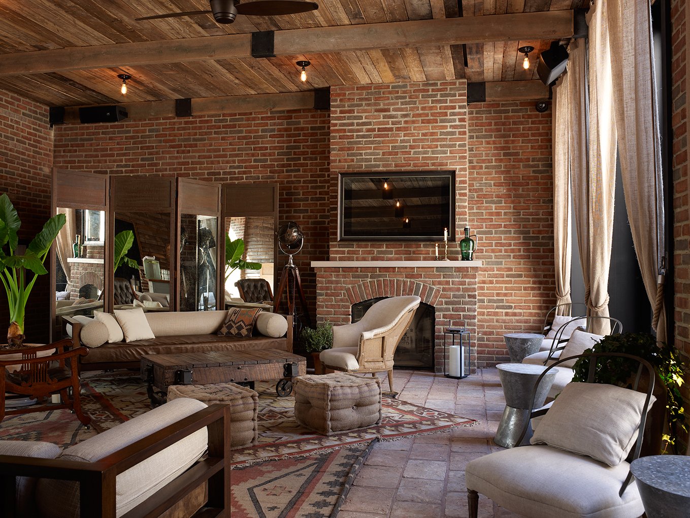 A rustic and cozy lounge features a seating area with various sofas and chairs, a low wooden coffee table on casters, and a fireplace set into a prominent brick wall, all beneath a slatted wood ceiling.