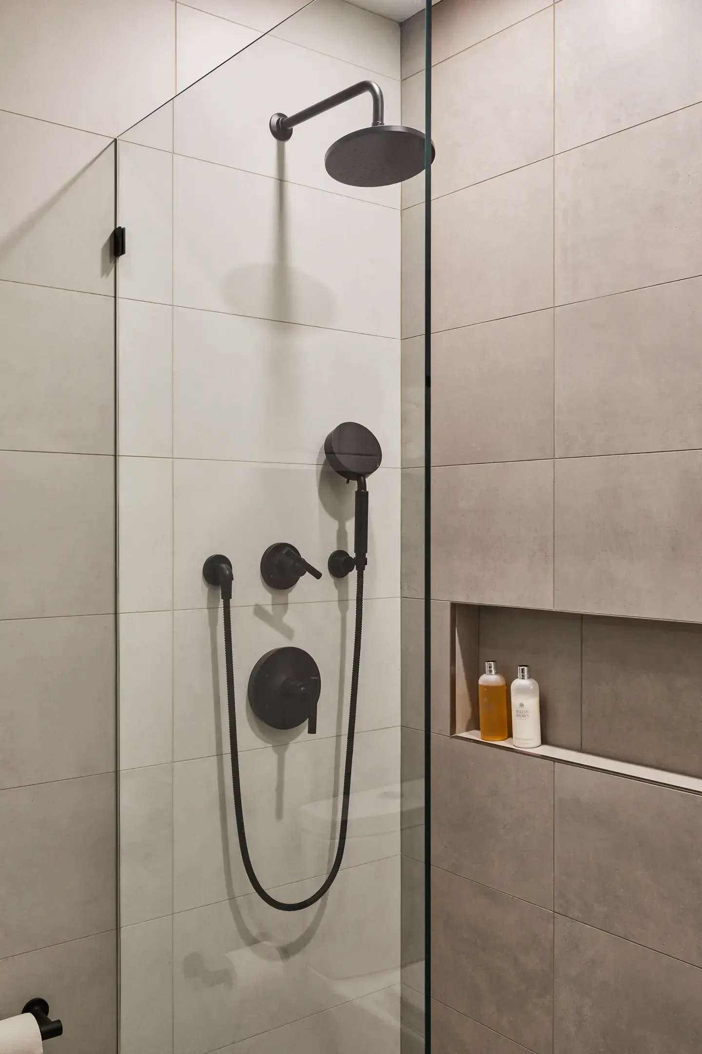 Modern walk-in shower with large-format neutral tiles, a matte black wall-mounted rain showerhead, matching handheld sprayer, minimalist controls, clear glass partition, and a recessed wall niche that enhances both function and clean design.