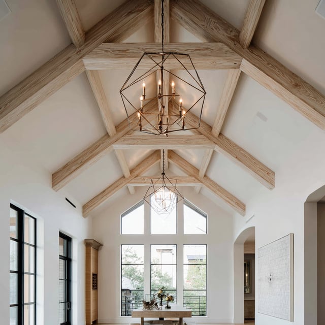 A bright vaulted room with exposed wooden ceiling beams, geometric chandeliers, and tall windows letting in natural light.