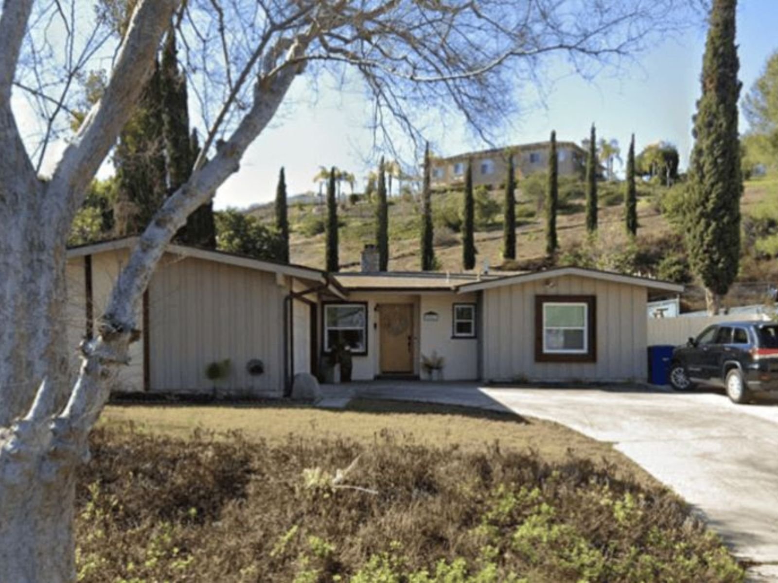A classic single-story ranch-style home featuring light beige board-and-batten siding, dark wood window trim, and a long concrete driveway set against a hillside lined with tall cypress trees.