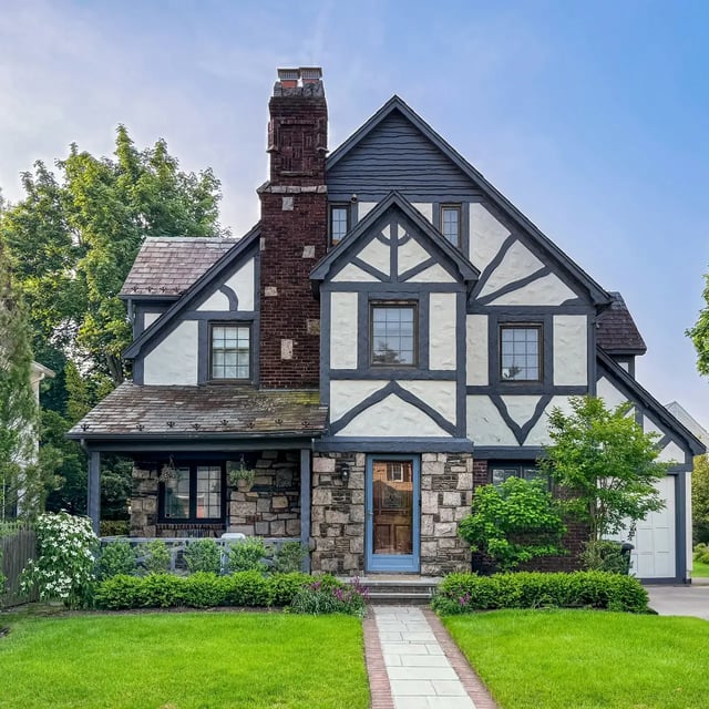 A two-story Tudor-style house with dark gray trim, white stucco, and a stone and brick chimney is shown, surrounded by a green lawn and landscaping.