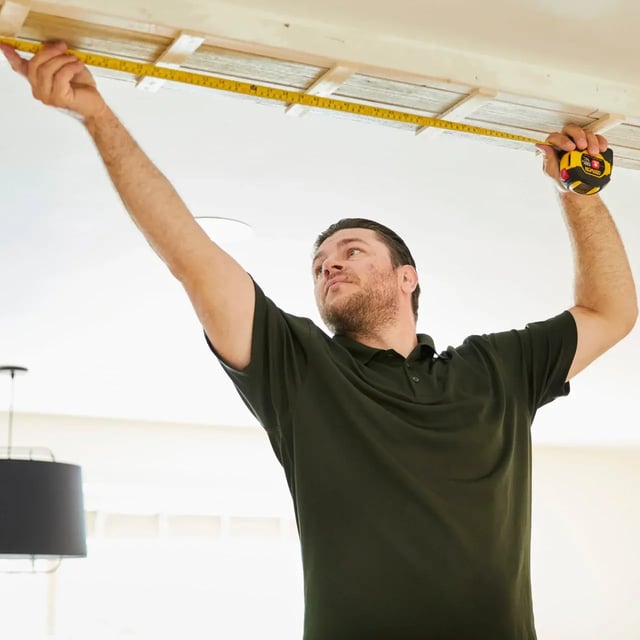  Man in a green shirt measures a ceiling with a tape ruler.