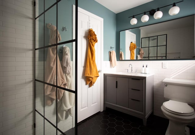 A modern bathroom with teal walls features a black-framed glass shower, white subway tile, a dark vanity with an undermount sink, a large rectangular mirror, black hex tile flooring, and mustard and beige towels hanging on hooks and racks.
