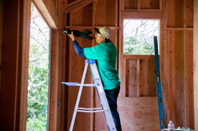 A person wearing a green shirt and cap stands on a step ladder inside a partially framed wooden building, using a power drill to work near the top of a door. Unfinished walls, exposed studs, and a level leaning against the wall are visible, with trees seen through the windows.