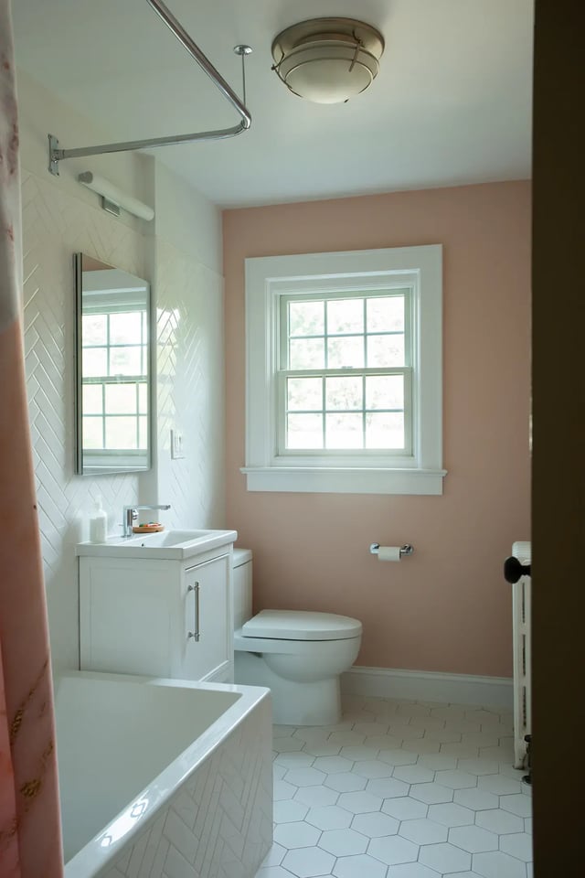 A bathroom featuring a white vanity, toilet, and bathtub, with light pink walls and white herringbone and hexagonal tiles.
