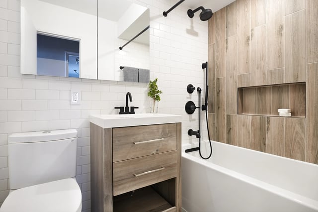 A modern bathroom features a white subway tile wall, a light wood-look vanity with a white countertop and a vessel sink with black fixtures, a toilet on the left, and a white bathtub with black shower fixtures and a wood-look tiled shower wall with a recessed niche.