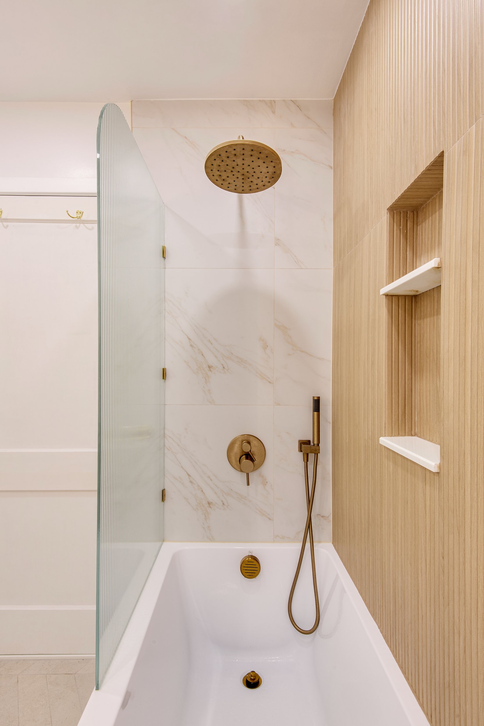 Modern tub-shower combo with a ceiling-mounted rainfall showerhead, handheld sprayer, fluted glass panel, marble-look wall tile, and built-in niche for a refined, space-saving shower upgrade.