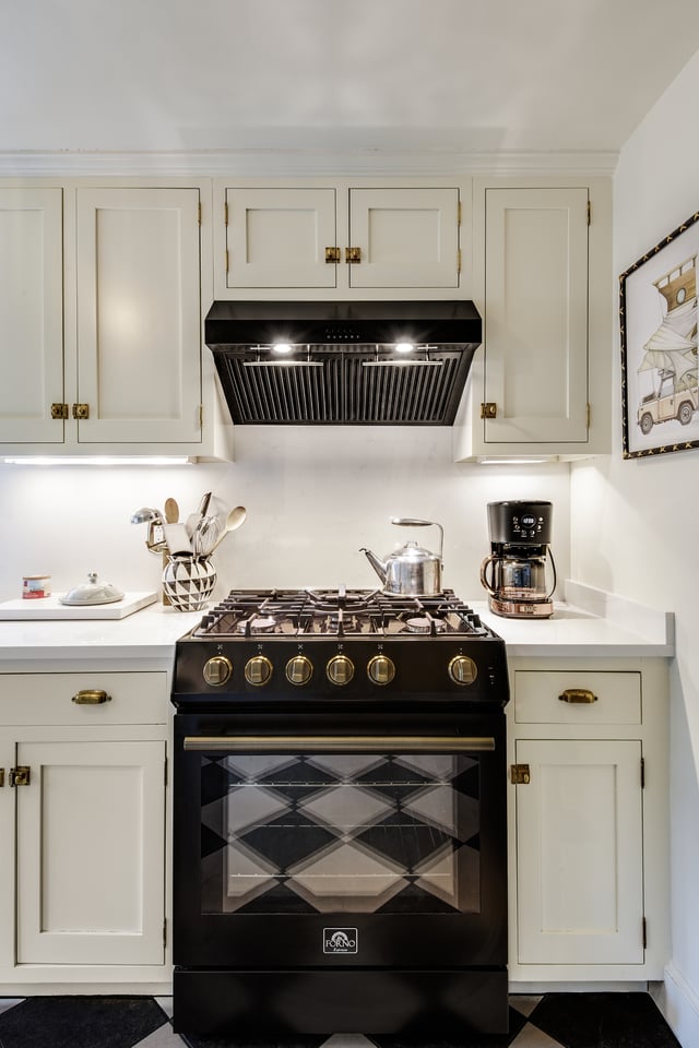 A black freestanding gas range with brass knobs is centered beneath a black vent hood, surrounded by white cabinetry and countertops with kitchen utensils, a kettle, and a coffee maker.