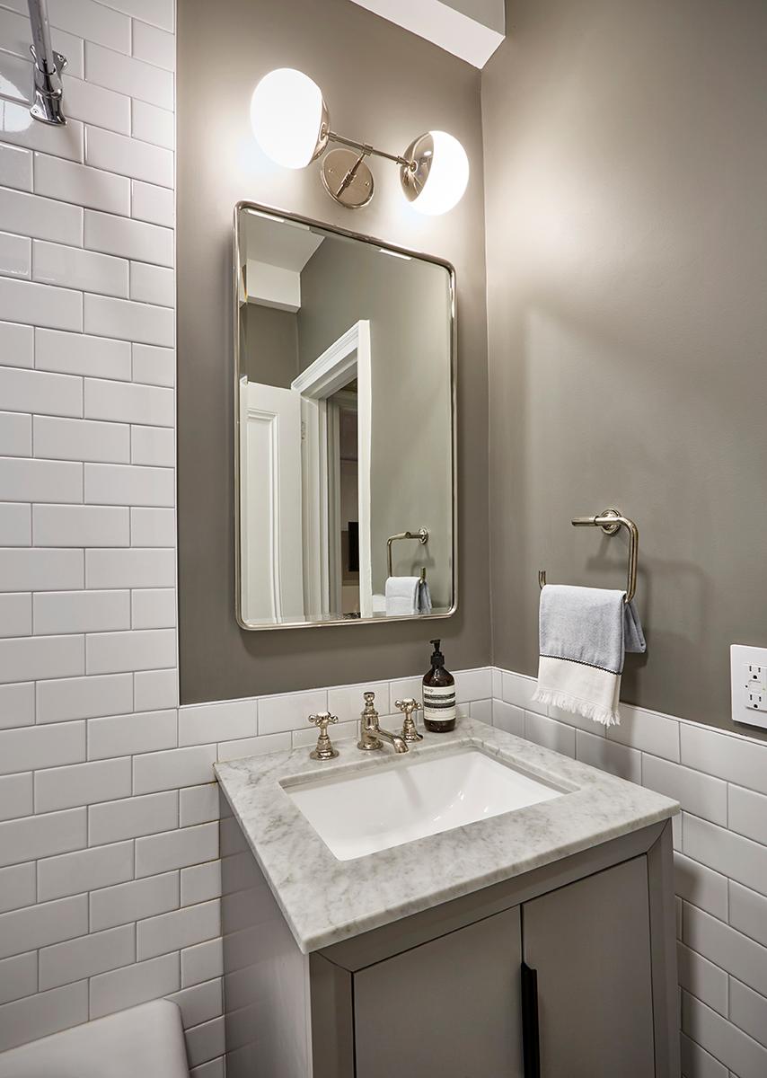 A modern bathroom vanity with a white rectangular sink and a marble countertop sits against a wall covered in white subway tiles on the lower half and gray paint on the upper half. Above the sink is a mirrored cabinet with a silver frame and a double-globe vanity light. A silver towel ring with a white and blue towel hangs on the right.