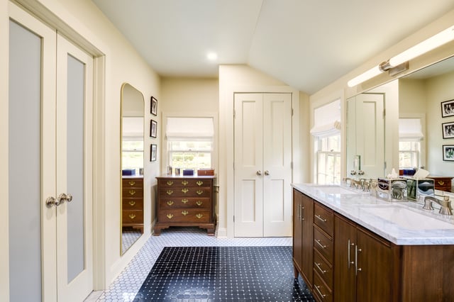 A brightly lit bathroom featuring a dark wood double vanity with a white marble countertop, a large mirror, two windows, and a dark tile floor with a contrasting white hexagon pattern border.