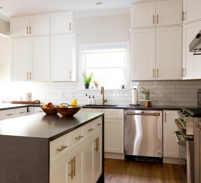 A modern kitchen featuring white Shaker-style cabinetry with gold hardware, a dark gray quartz island with a waterfall edge, and a classic white subway tile backsplash.