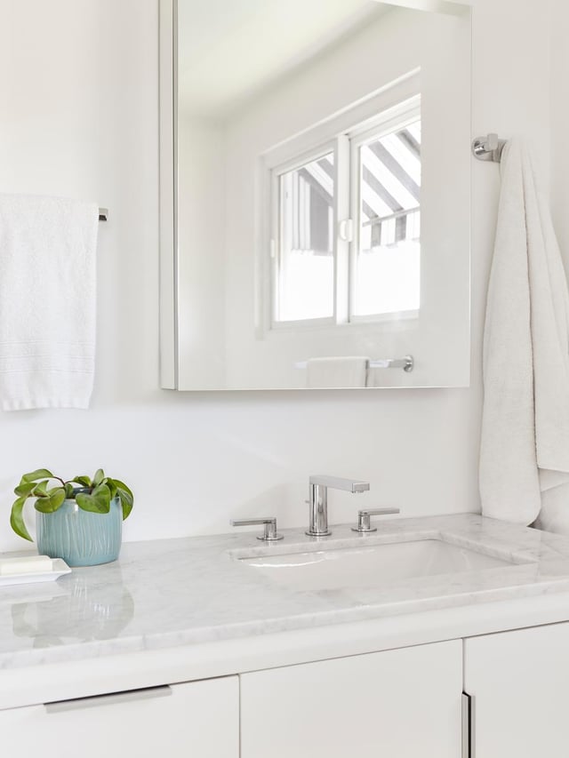 A bright, modern bathroom vanity with a white marble countertop, built-in sink, chrome faucet, wall-mounted mirror reflecting a window, white towels on chrome hooks, and a small potted plant for accent.