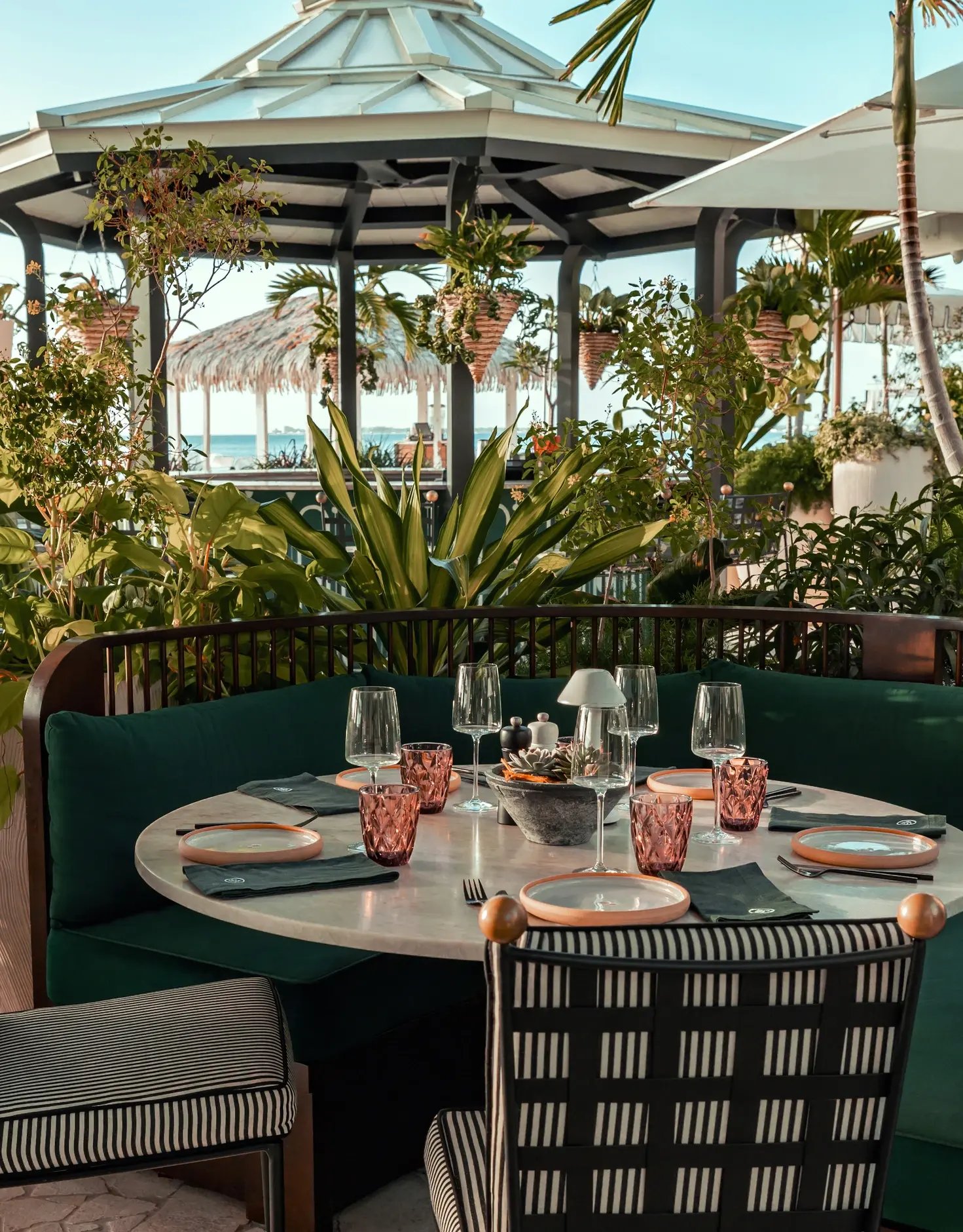 An outdoor dining setting features a round white marble table set for a meal with pink glassware, surrounded by a deep green upholstered banquette and black and white patterned chairs, all nestled among lush tropical plants beneath a white gazebo.