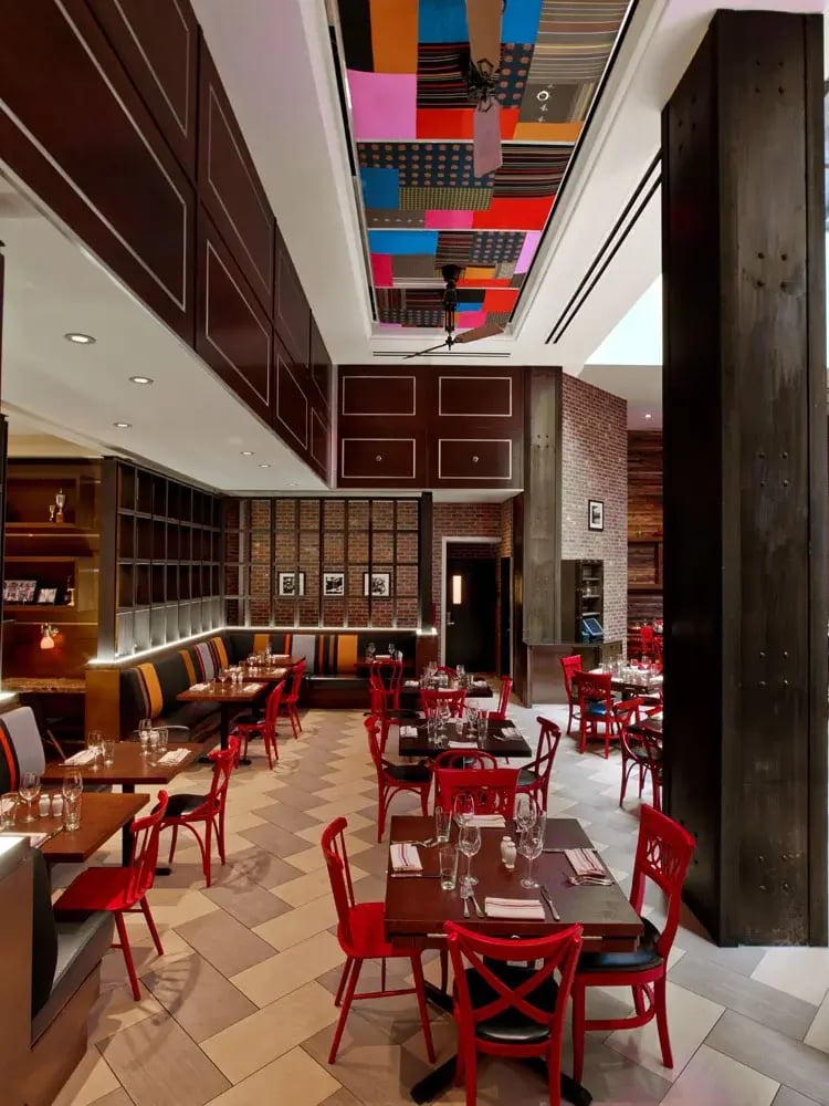An eclectic restaurant interior features tables with bright red chairs, banquette seating along a brick and dark wood paneled wall, a floor with a geometric pattern of tan and white tiles, and a colorful patchwork ceiling with a patterned design.