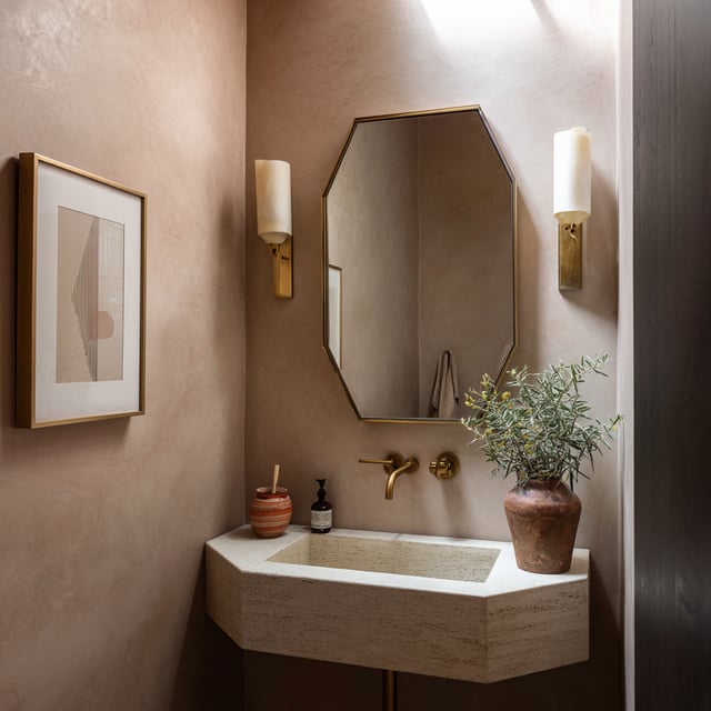 A powder room features smooth, dusty-rose-colored plaster walls, an octagonal gold-framed mirror flanked by two cylindrical sconces, a small beige stone sink, and a framed piece of abstract art.
