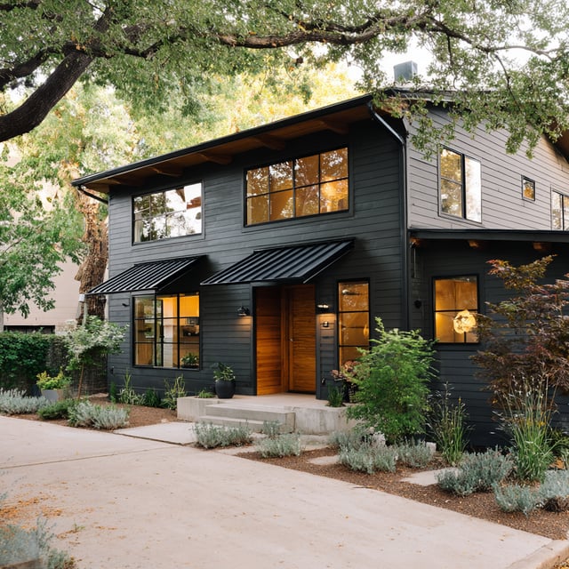 A modern, two-story house with dark gray siding, black accents, and a wood front door, surrounded by lush greenery.