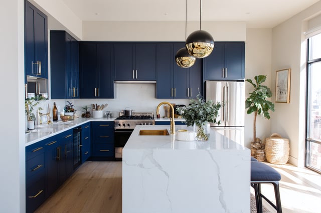 A modern kitchen features navy blue cabinets with gold-colored hardware, a large white marble waterfall island, and gold-colored pendant lights, with a stainless steel refrigerator and stove visible.