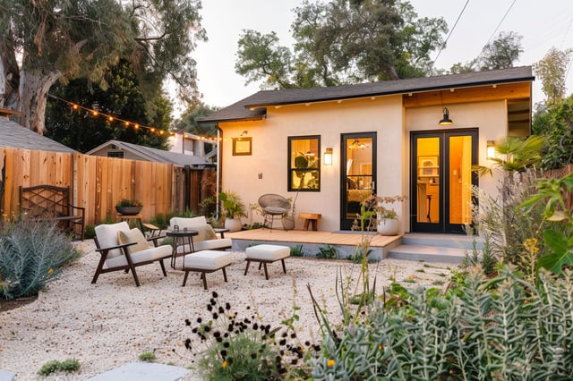 A cozy and inviting Accessory Dwelling Unit (ADU) at dusk, featuring warm interior lighting, a small wooden deck, and a minimalist gravel courtyard.