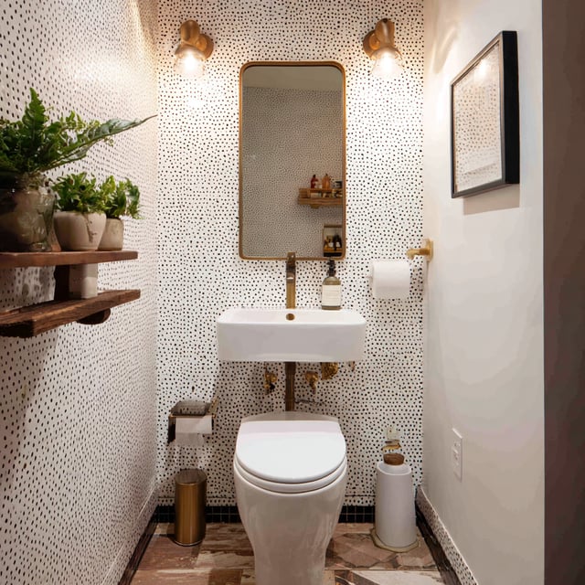 Small powder room featuring a wall-mounted white sink and toilet, dotted wallpaper, brass fixtures, a rounded mirror with sconces, wooden shelves with plants, and a patterned stone floor.